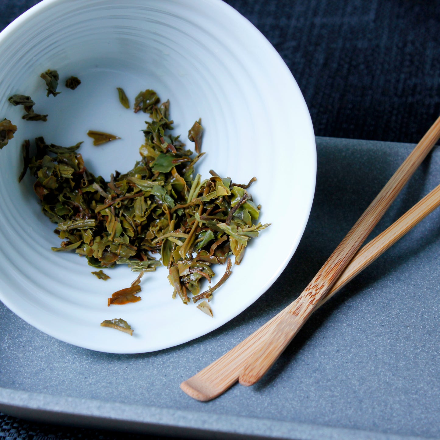 Darjeeling 1st Flush: Mim Estate - wet leaves in a white bowl, two tea rakes next to is on a grey tray. Tasting Notes:
grapefruit • coriander • cedar