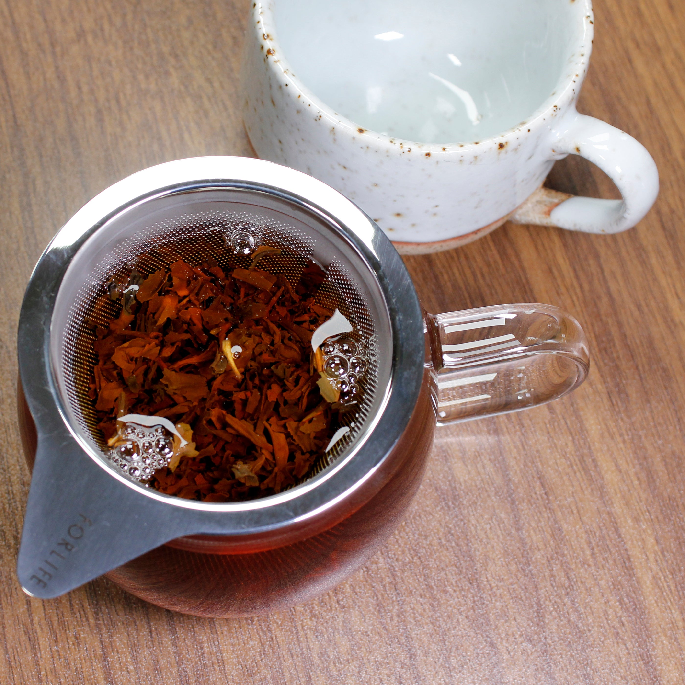 down shot of the tea in the mug with tea leaves in the strainer. A white and brown speckled mug is next to it.