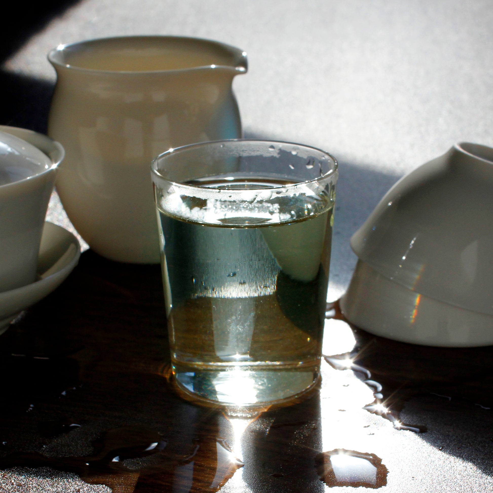 Glass of tea surrounded by cups, a pitcher and Gaiwan on a dark wood table. Tasting Notes:
honeysuckle • macadamia • alfalfa