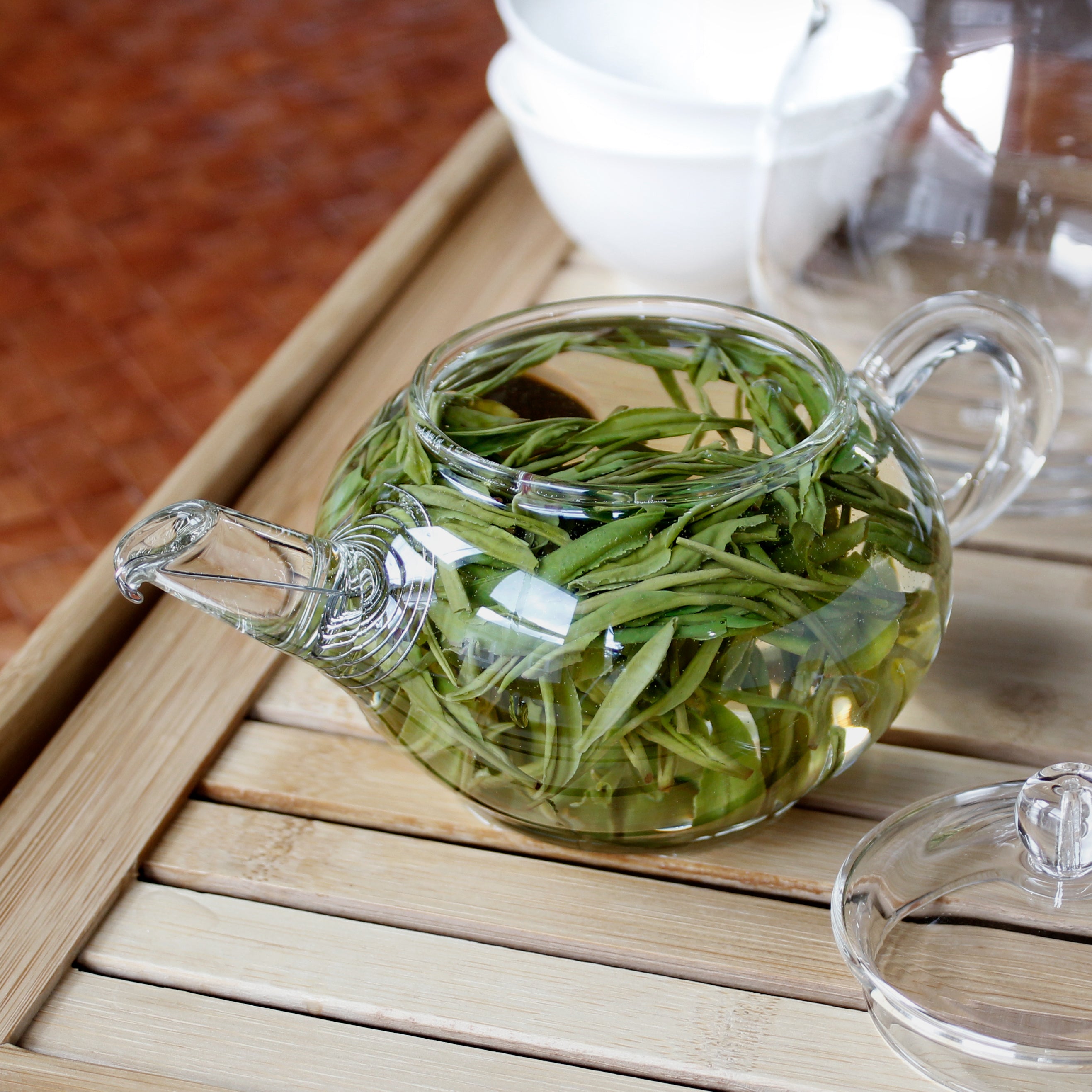 Clear glass teapot filled with green tea leaves on a wooden surface. White tasting cups and pitcher in the background. Tasting Notes:
honeysuckle • macadamia • alfalfa