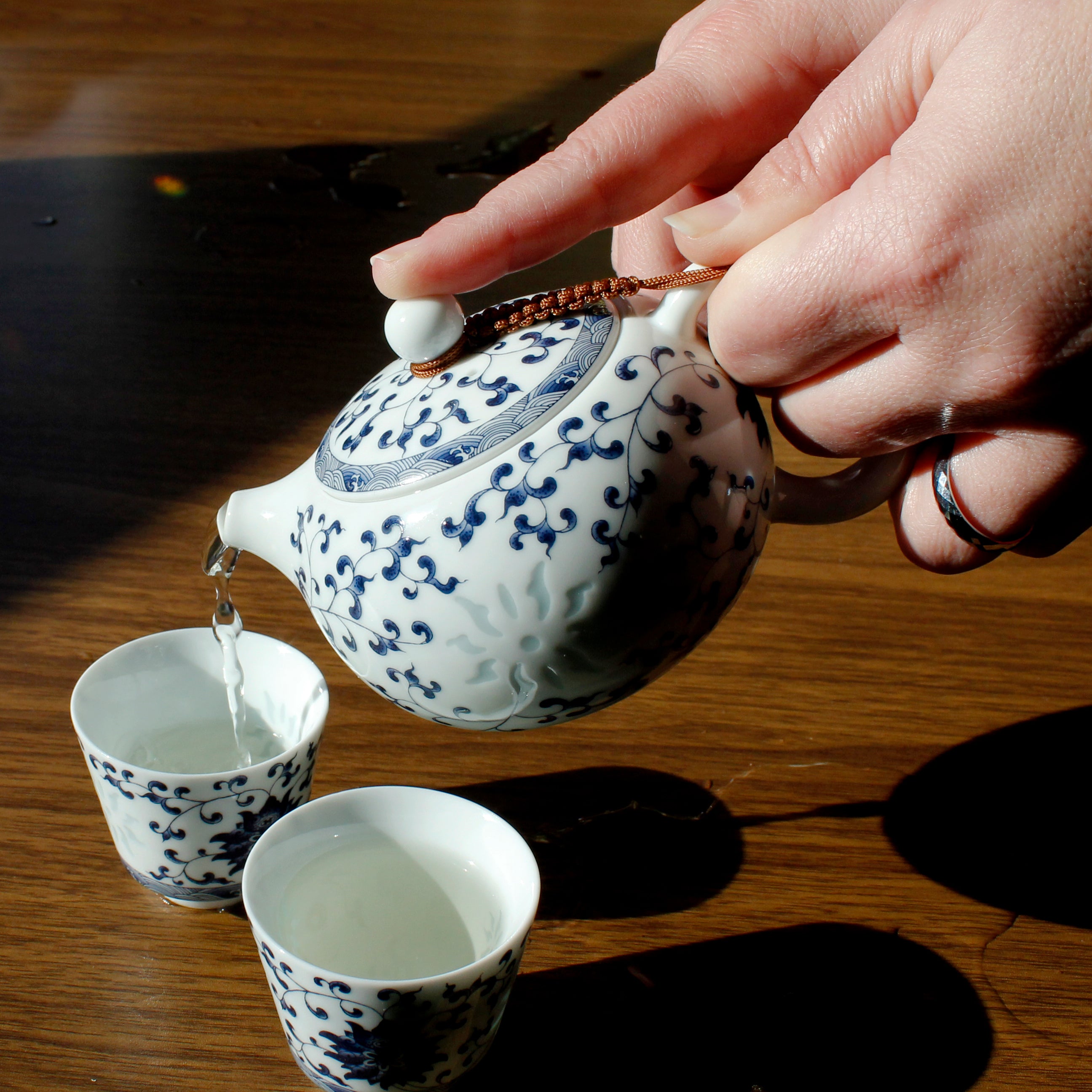 Person pouring tea from a blue and white teapot into the matching cups on a wooden surface.
