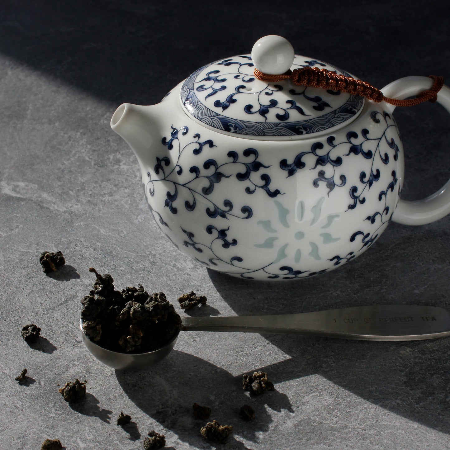 White teapot with blue floral patterns on a dark surface, surrounded by tea leaves and a tea scoop.