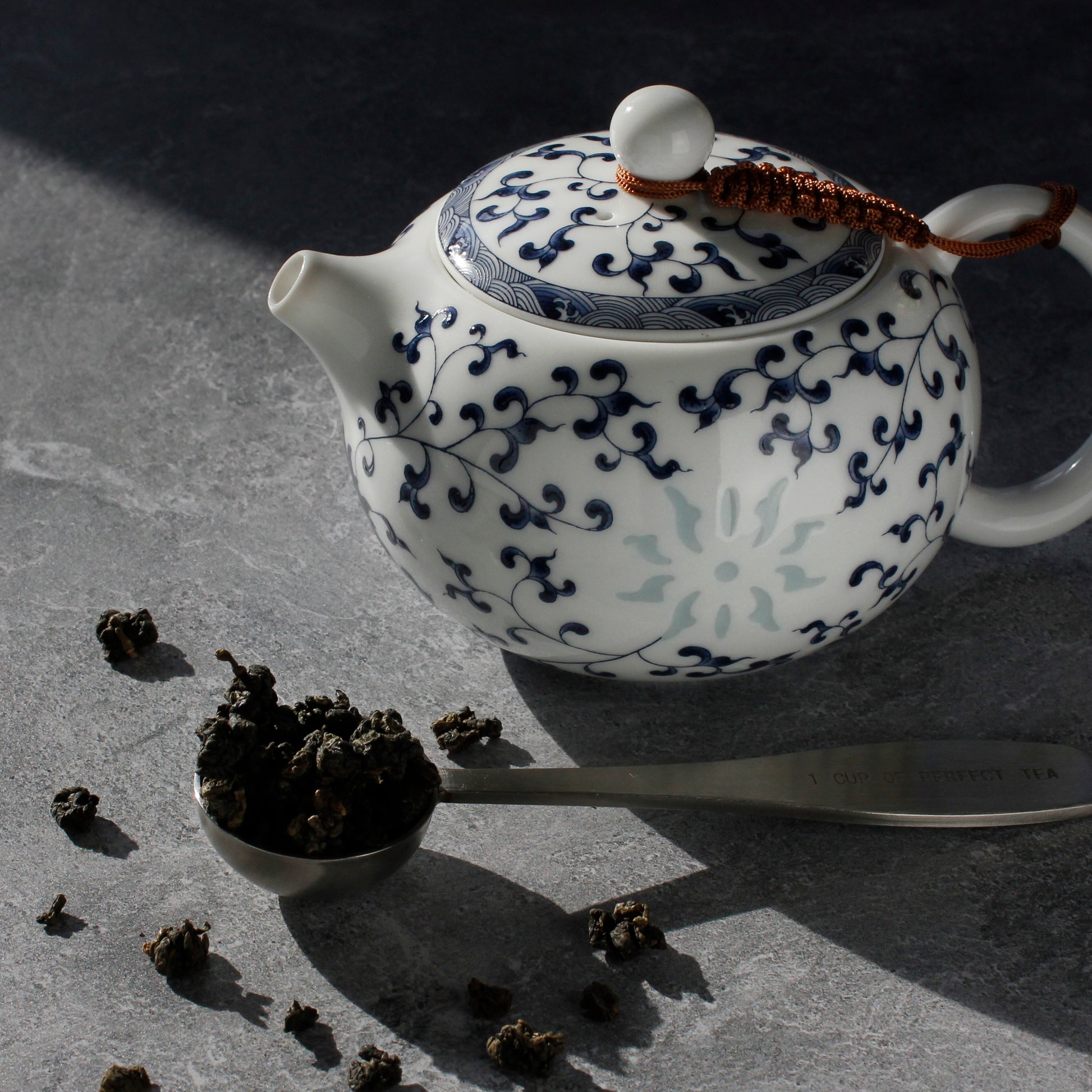 White teapot with blue floral patterns on a dark surface, surrounded by tea leaves and a tea scoop.