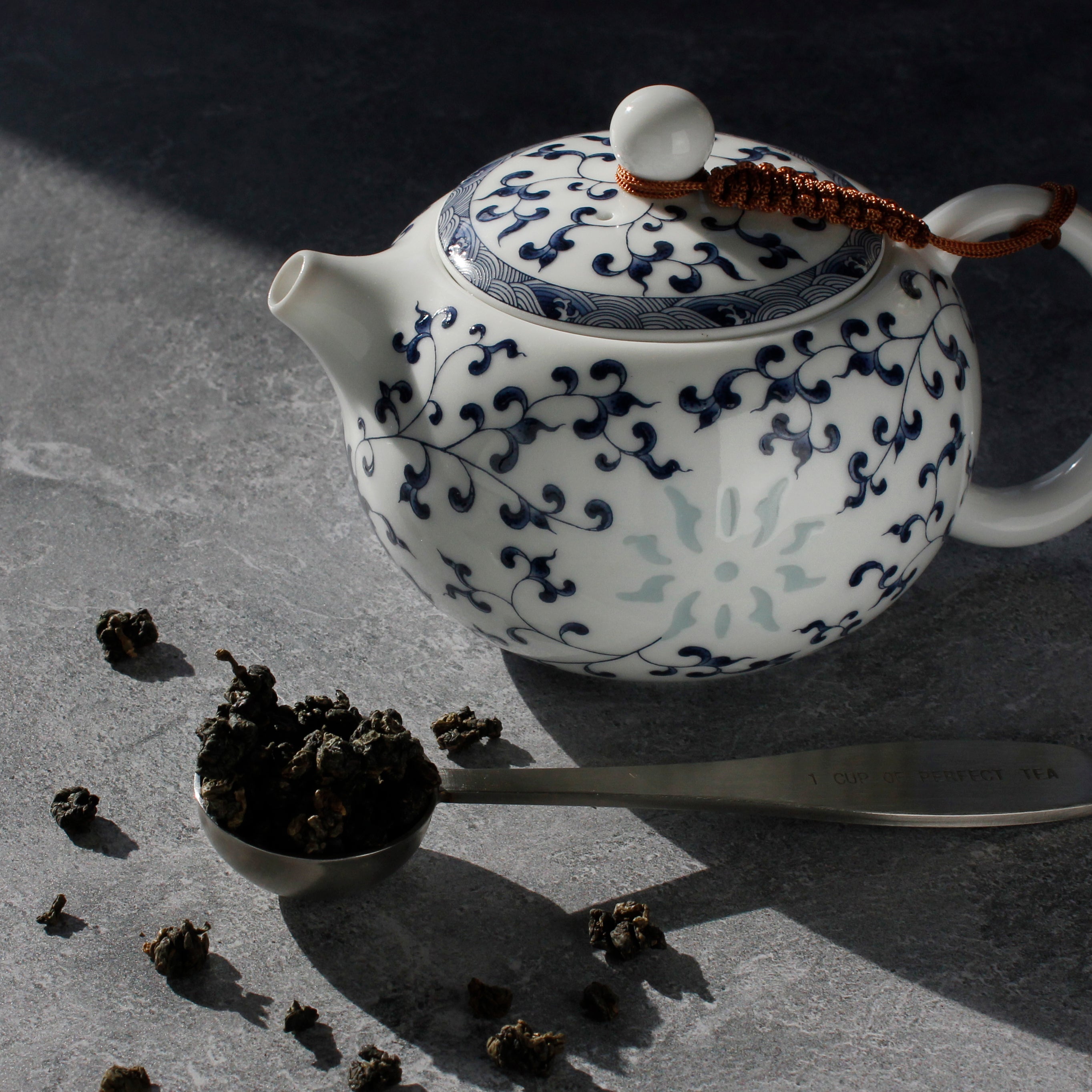 White teapot with blue floral patterns on a dark surface, surrounded by tea leaves and a tea scoop.