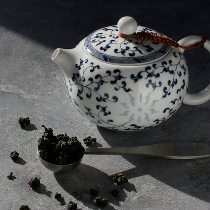 White teapot with blue floral patterns on a dark surface, surrounded by tea leaves and a tea scoop.
