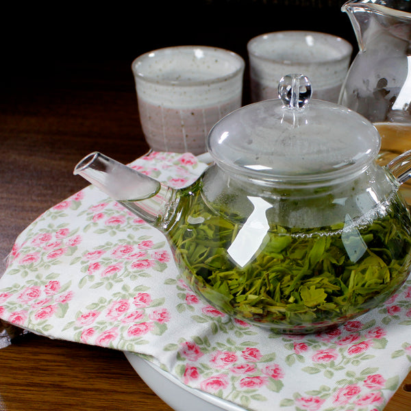 The beautiful Clouds & Mist green tea steeping in a glass teapot, on a pink tea towel, 2 cups and a pitcher in the background.