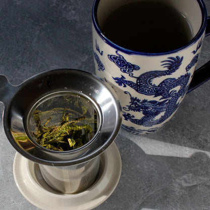 The tea strainer with tea leaves on the lid next to  blue and white dragon mug on a gray surface