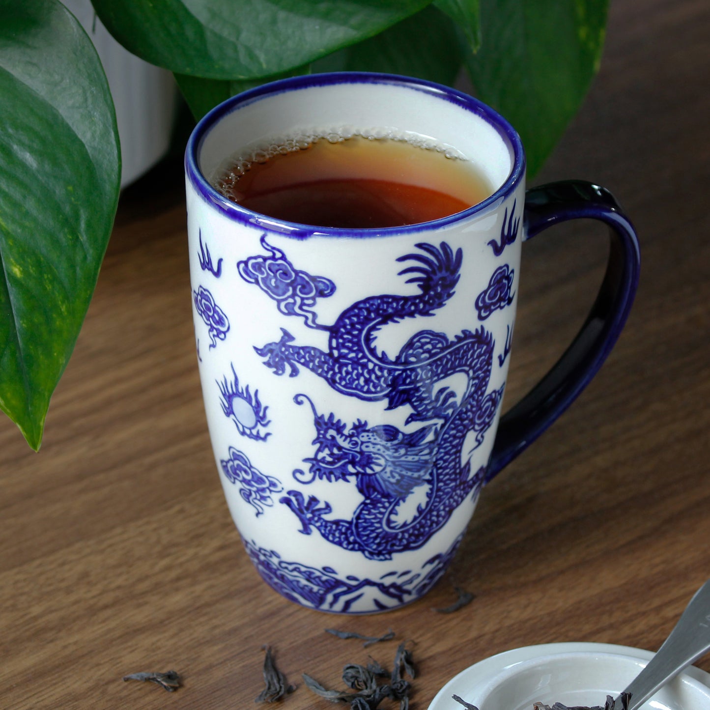 Blue and white ceramic mug with dragon design on a wooden table, filled with tea. Plants in the background and small bit of dried tea and scoop can be seen in the foreground. 