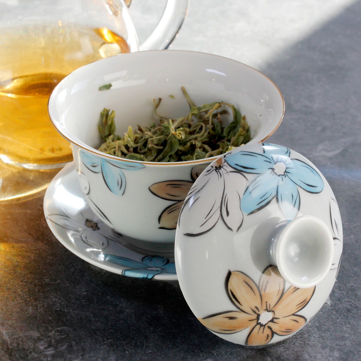 Gaiwan with floral design on a saucer, lid to the side to show the wet tea leaves inside, brewed tea in glass pitcher to the side on a grey background. 