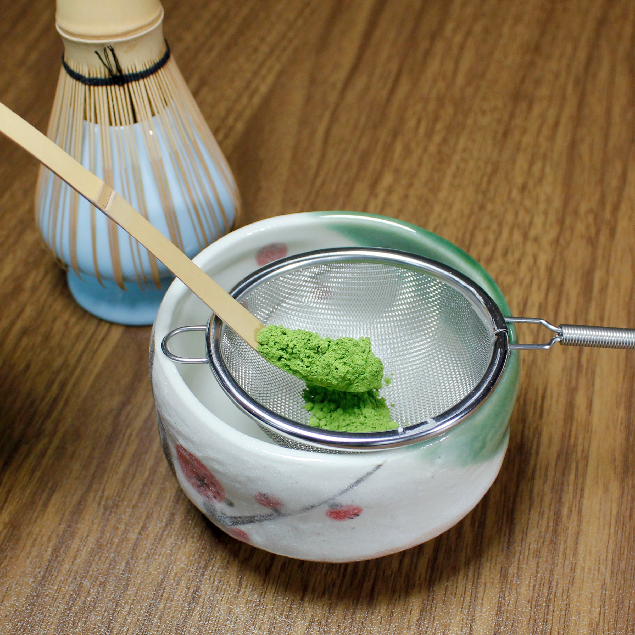 Matcha scoop holding matcha over a sifter resting on a matcha bowl. Behind is a bambo match whisk it its chasen holder. The whole set in on wood table.