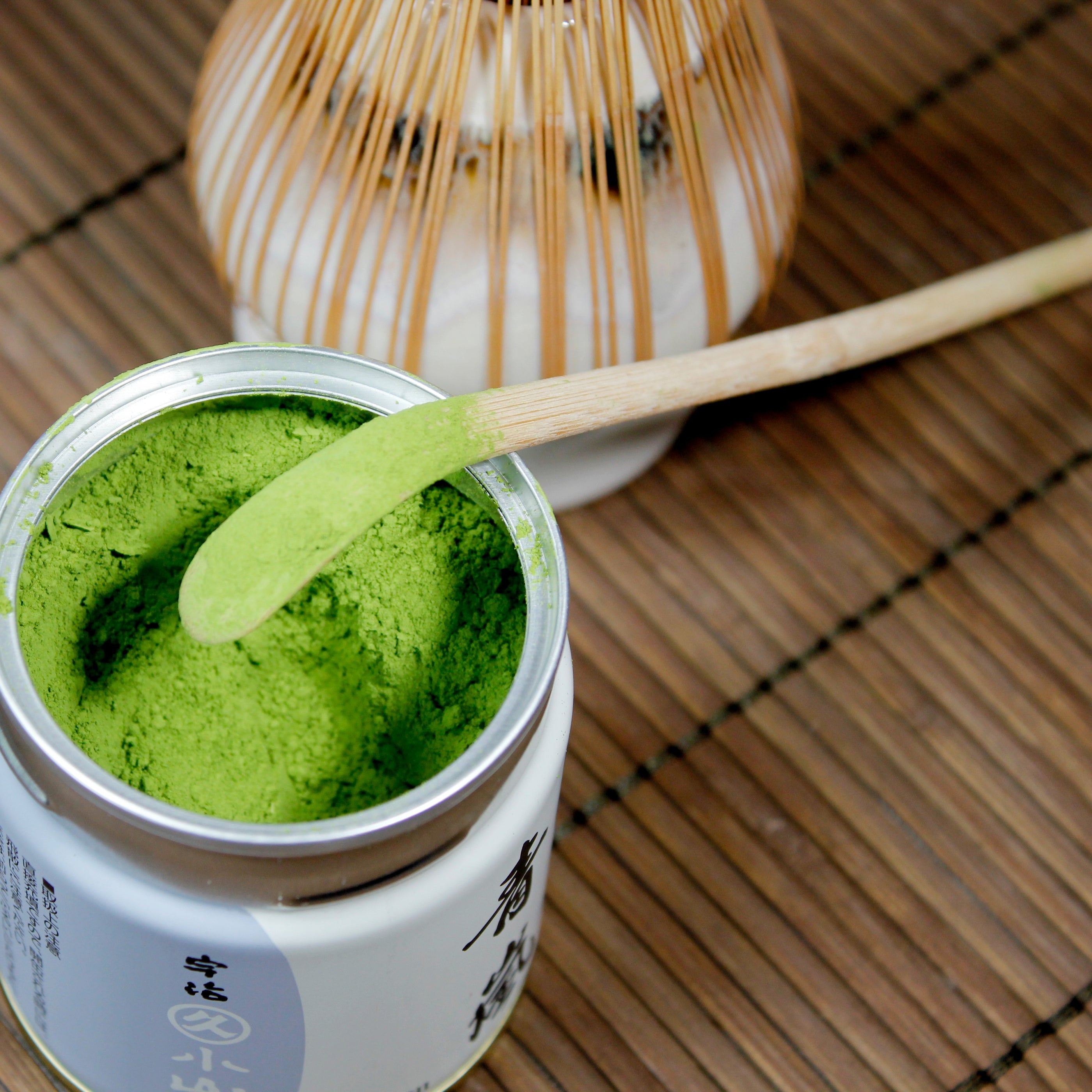 Open container with matcha, the scoop is laying on the open lip. Matcha whisk and chasen in the background.