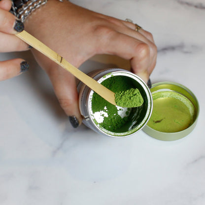 Person scooping green matcha powder from a container with a wooden spoon.
