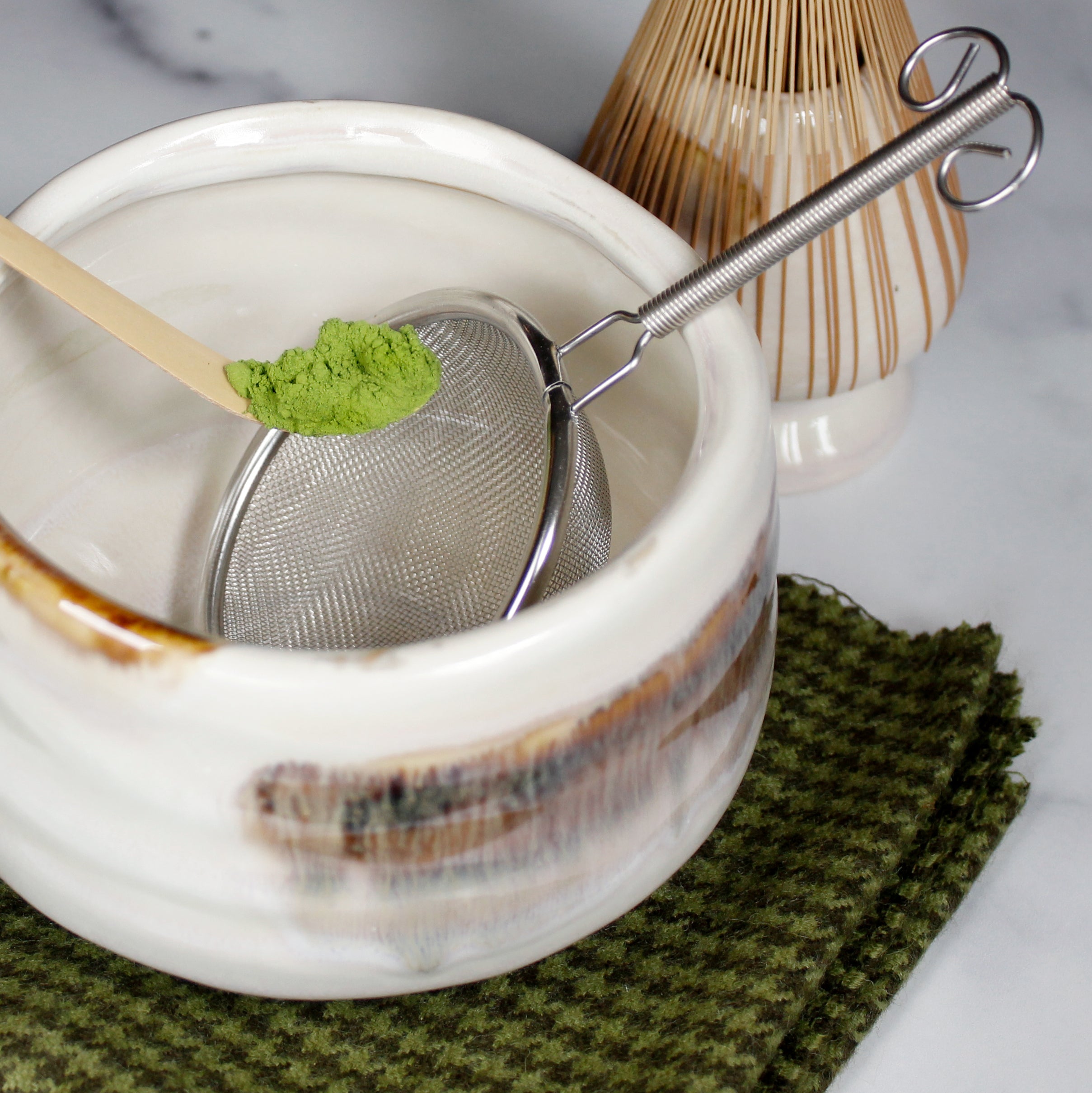 The sifter sitting in a Matcha bowl as a Matcha scoop with Matcha hovers over it.