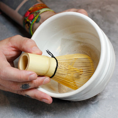 Hands holding a matcha whisk and off white matcha bowl at an angle too see inside. 