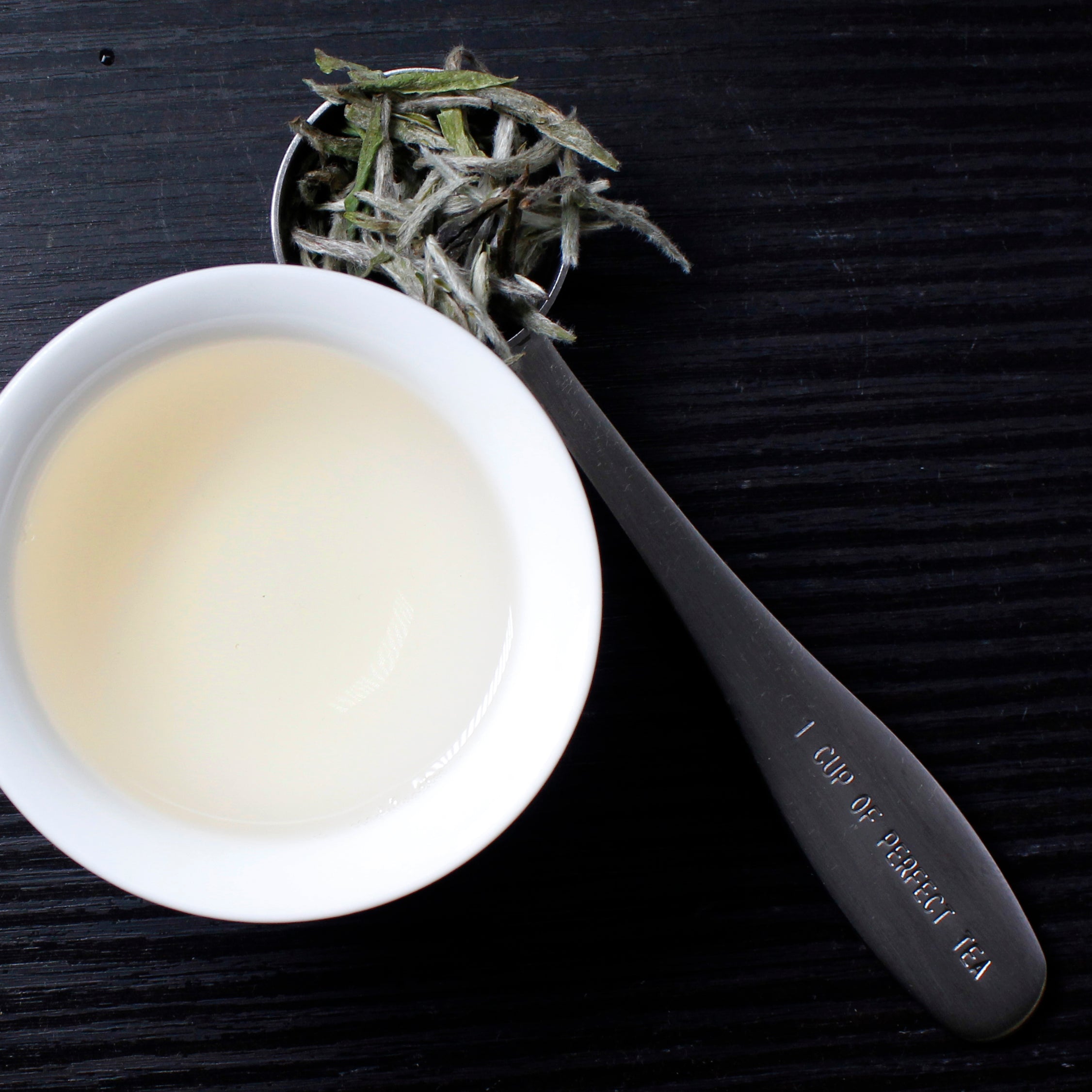 Tea scoop filled with soft tea leaves next to the cup with brewed tea on a black wood background.