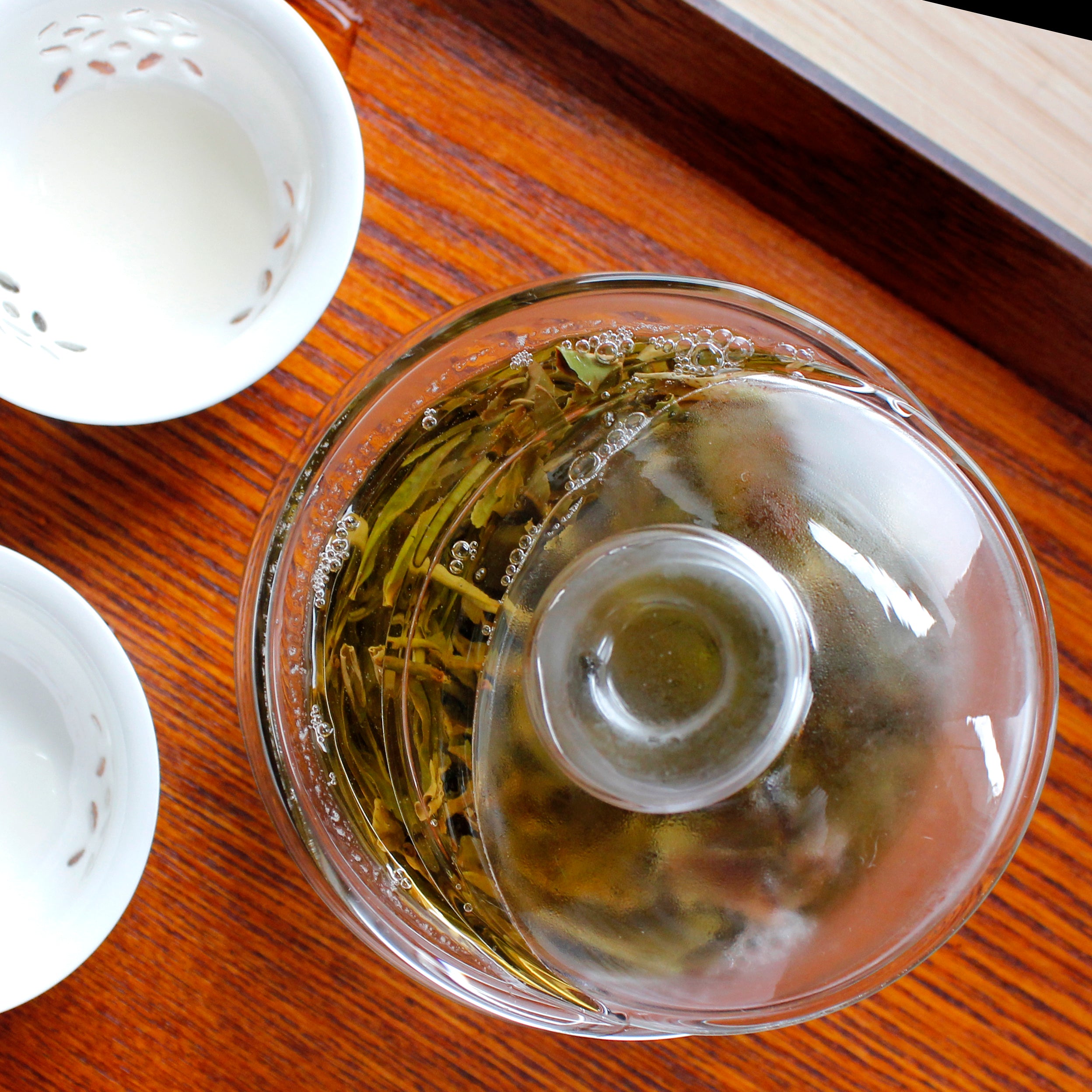 Downward shot of filled gaiwan, hot steam clouding the glass. Two cups are next to the gaiwan, a wood table underneath. 