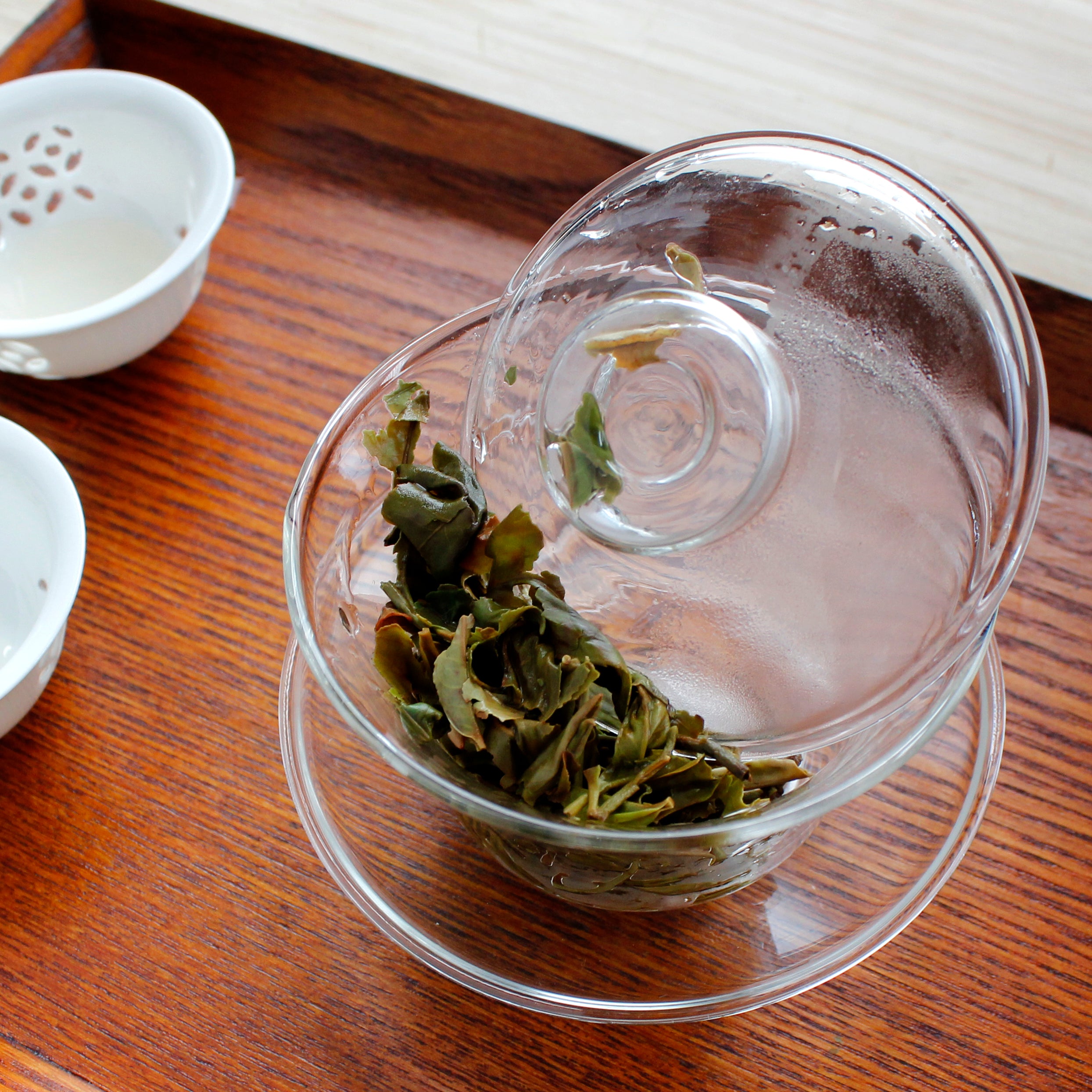 Gaiwan with wet tea leaves pushed to the side, two small cups are next to it, on a wood table. 