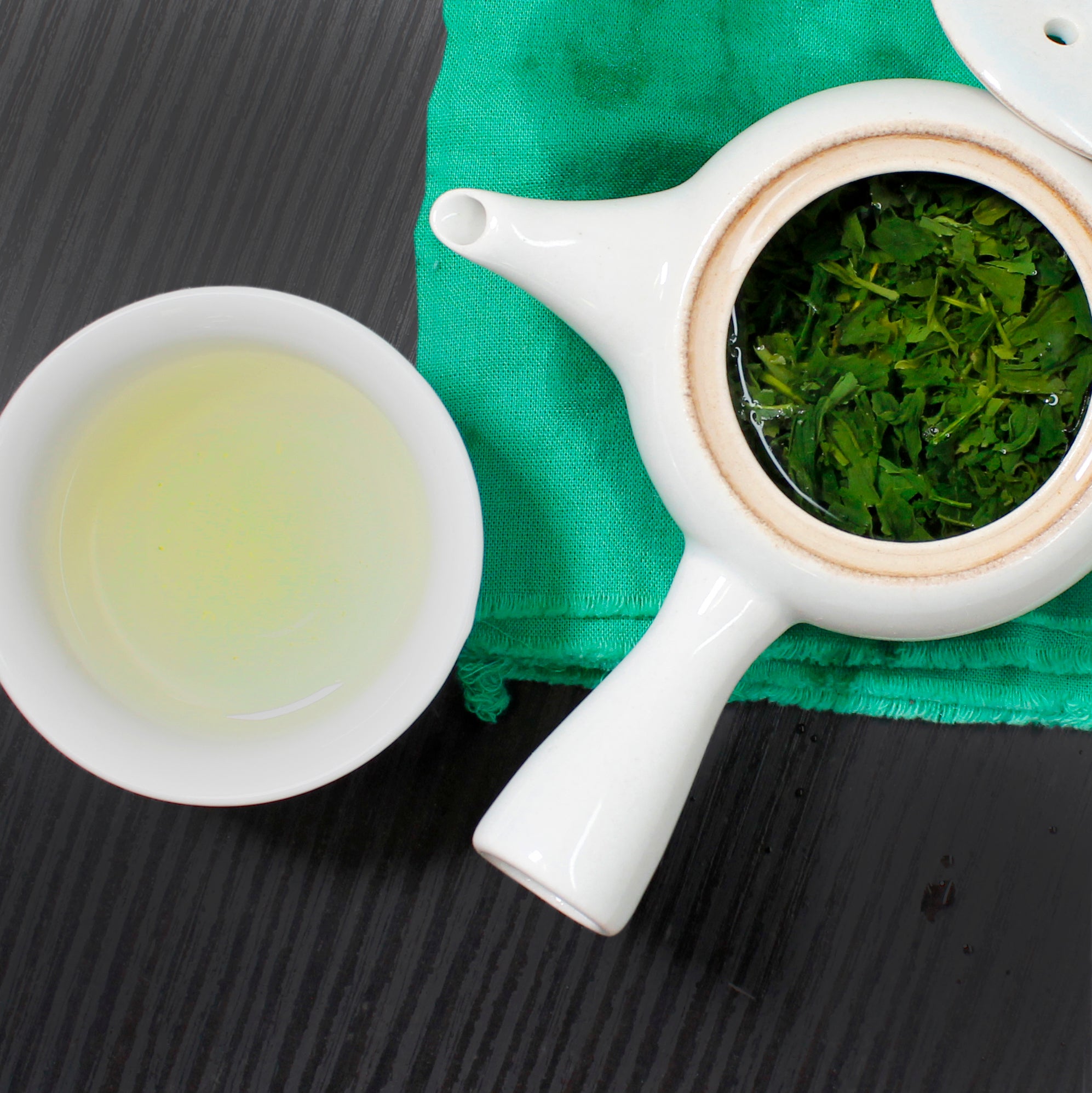 White tea pot with handle on a green tea towel,  lid is off to show tea and next to it is a tea cup with tea. 