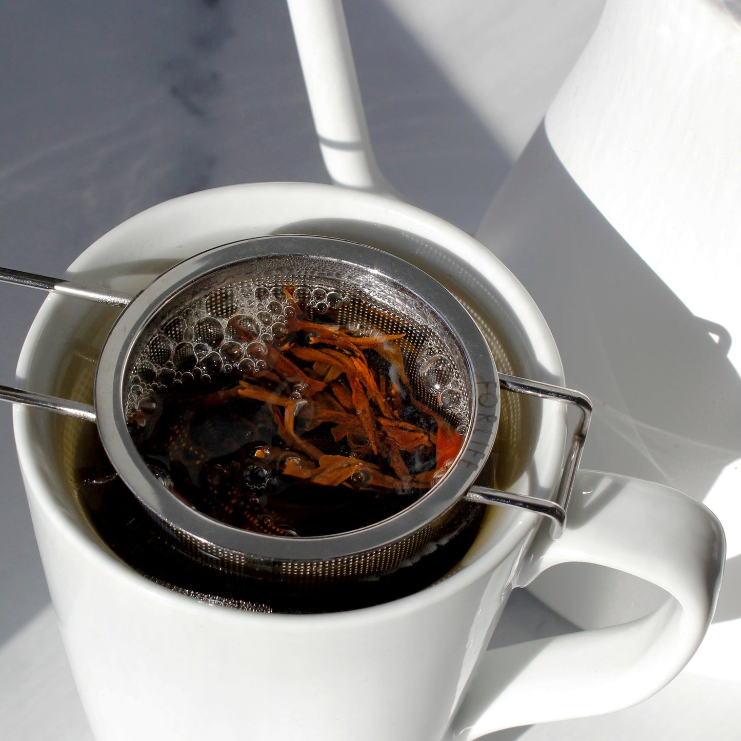 White mug with metal tea infuser filled with tea leaves on a white background