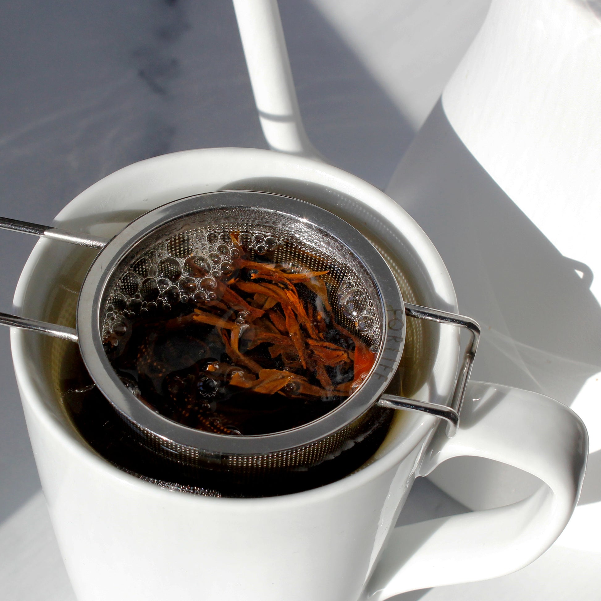 White mug with metal tea infuser filled with tea leaves on a white background